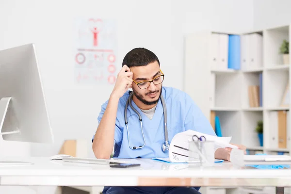 Portrait of young Middle-Eastern doctor wearing glasses sitting at desk in office and reading patients reports