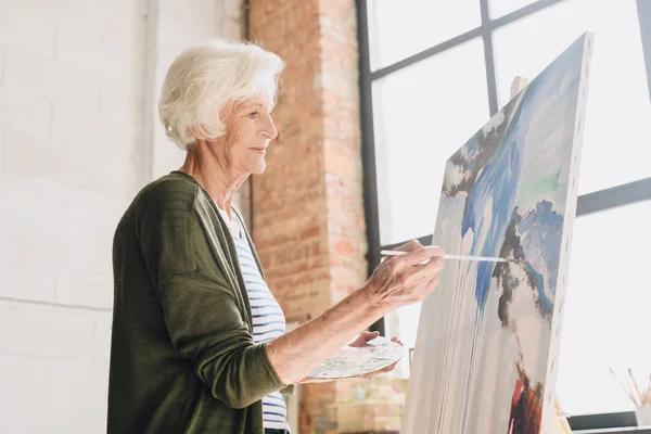 白い髪の年配の女性の絵を描くイーゼル アート スタジオに立ち向かう日光の下で Windows でパレットを保持している像側ビュー — ストック写真