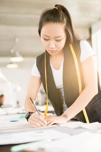 Waist up portrait of focused Asian woman drawing fashion sketches while standing at tailors table and working in modern design atelier lit by sunlight