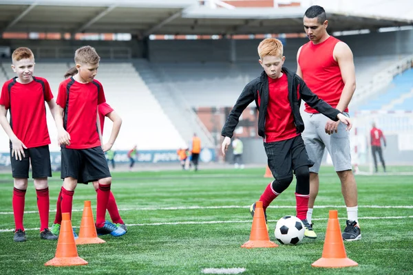 Full length portrait of junior football team practicing in stadium, row of teenage boys leading ball between orange cones, copy space