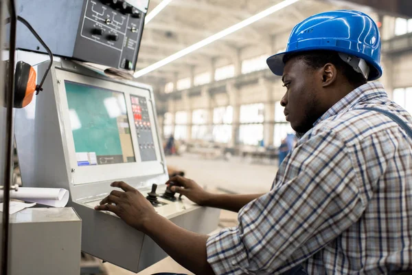 Profile view of highly professional worker wearing protective helmet and overall operating machine at production department of modern plant, portrait shot