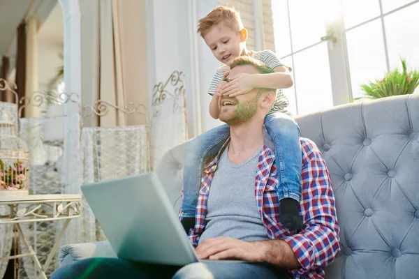 彼の肩の上に座って 日光の家でプレイしながらノート パソコンを使用しているハンサムなお父さんの目を覆っているかわいい男の子の肖像画 — ストック写真