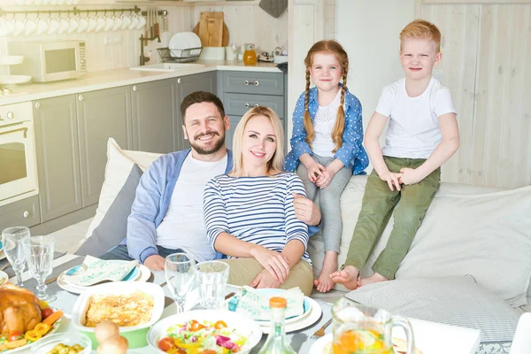 Portrait of successful family enjoying dinner together in modern apartment posing on couch with two adorable children, all looking at camera