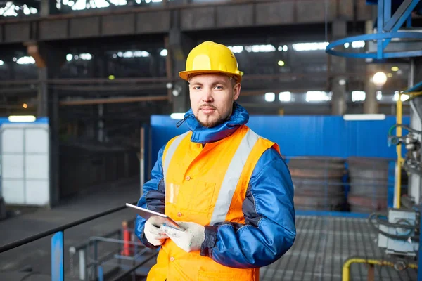 英俊的年轻工人穿制服和安全帽看着相机 同时手持数字平板电脑 以操作机器在现代工厂生产部门的腰向上的肖像 — 图库照片