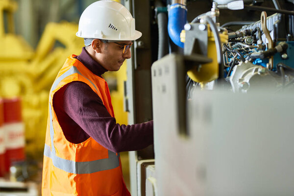 Profile view of mixed-race worker wearing reflective vest and protective helmet operating machine while standing at spacious production department of modern plant