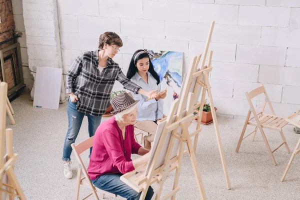 絵画クラスで若い女性にコメントを与える女性美術教師とイーゼルはアート スタジオで座っている学生の高角度の肖像画 — ストック写真