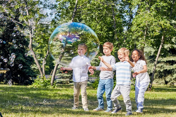 多民族儿童群体在被阳光照亮的公共公园里度过温暖的夏日时 与大肥皂泡一起享受乐趣 — 图库照片