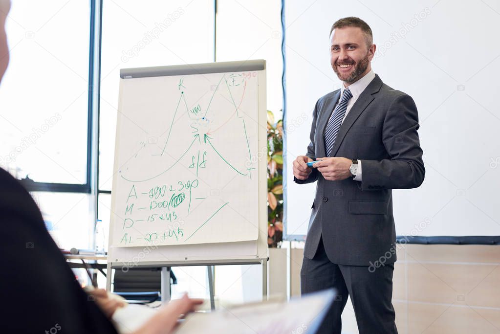 Portrait of bearded business coach smiling cheerfully while standing by whiteboard giving presentation for audience