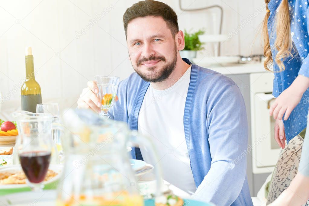 Portrait of smiling mature man looking at camera and holding glass of wine while enjoying family dinner at home in sunlight, copy space