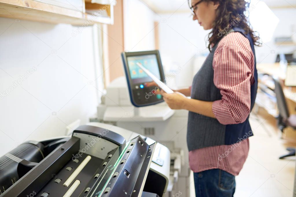 Side view of adult woman holding paper sheets and calibrating printer while working in nice printing office. 