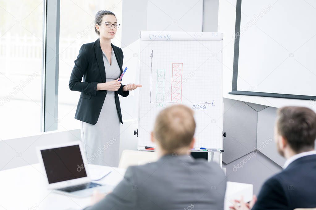 Portrait of young businesswoman wearing glasses standing at whiteboard and explaining marketing strategy  during meeting in conference room, copy space