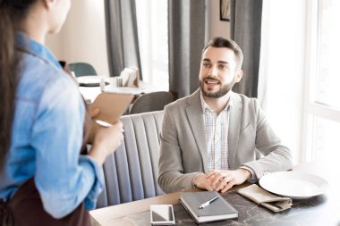 Neşeli yakışıklı genç işadamı rahat kıyafet ise sipariş verme ve günlüğünü ve restoranı smartphone ile masada oturan garson gülümseyerek sakallı