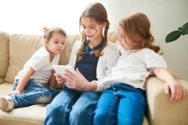 Rahat koltukta oturan ve o iç oturma odası arka plan üzerinde modern dijital tablet bilgisayar oyunu oynamak süre onların ablası eylemleri aşağıdaki iki küçük kız