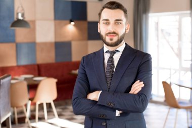 Sakal ve bıyık resmi ceket ve kollar göğüs üzerinde geçiş ve restoran kamerada bakarak kravat giyen olumlu yakışıklı genç işadamı içerik