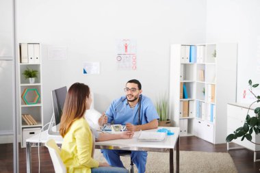 Ofiste masada oturan ve dikkatle bayan hasta için kan basıncı ölçme sırasında dinleme gözlüklü genç Orta Doğulu Doktor portresi geniş açı.