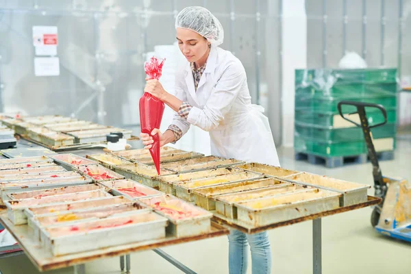 Confectionery factory employee in white coat squeezing red cream from pastry bag.