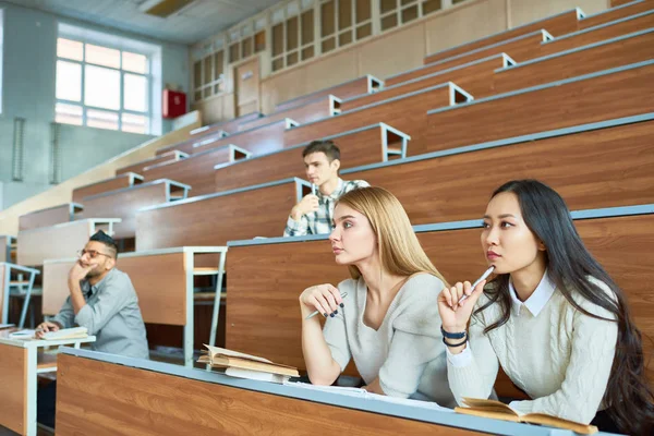 International group of people sitting at separate tables in lecture hall of modern college listening to professor, focus on two beautiful girls in foreground, copy space