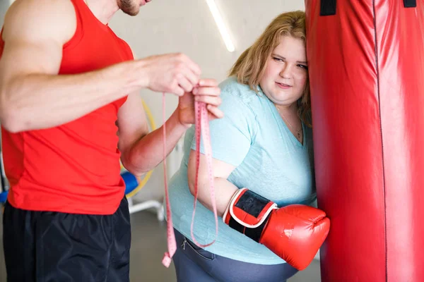 Portrait of exhausted obese woman looking at measuring tape during training in gym