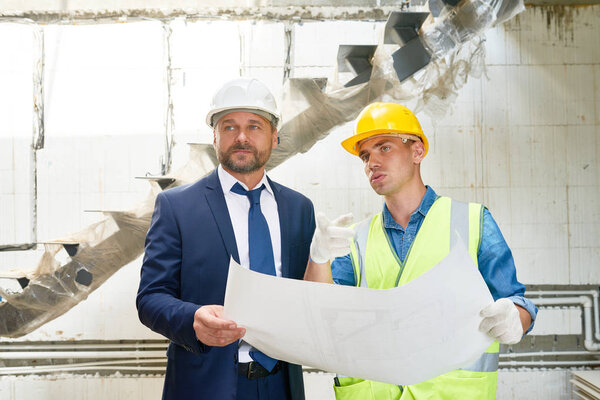 Waist up portrait of foreman and businessman wearing hardhat discussing floor plans on site standing by unfinished flight of stairs