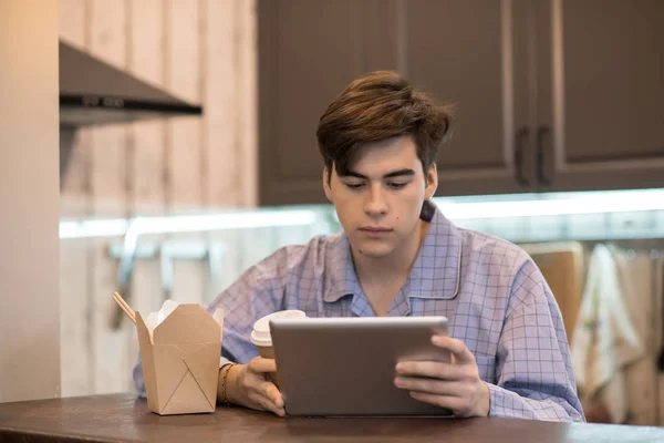 Young man in pajamas holding cup of hot beverage and browsing tablet while sitting at table near takeout box.