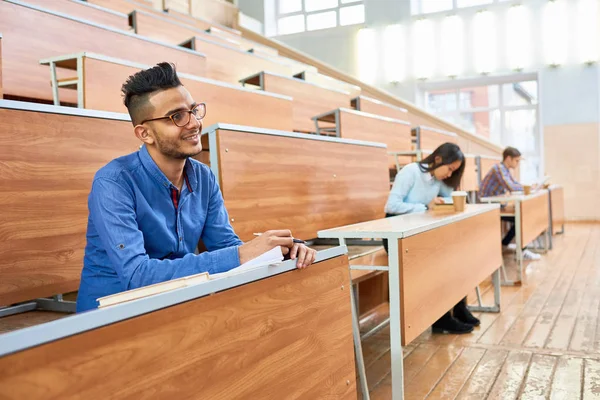 International group of people sitting at separate tables in lecture hall of modern college, focus on young Middle-Eastern man smiling in foreground