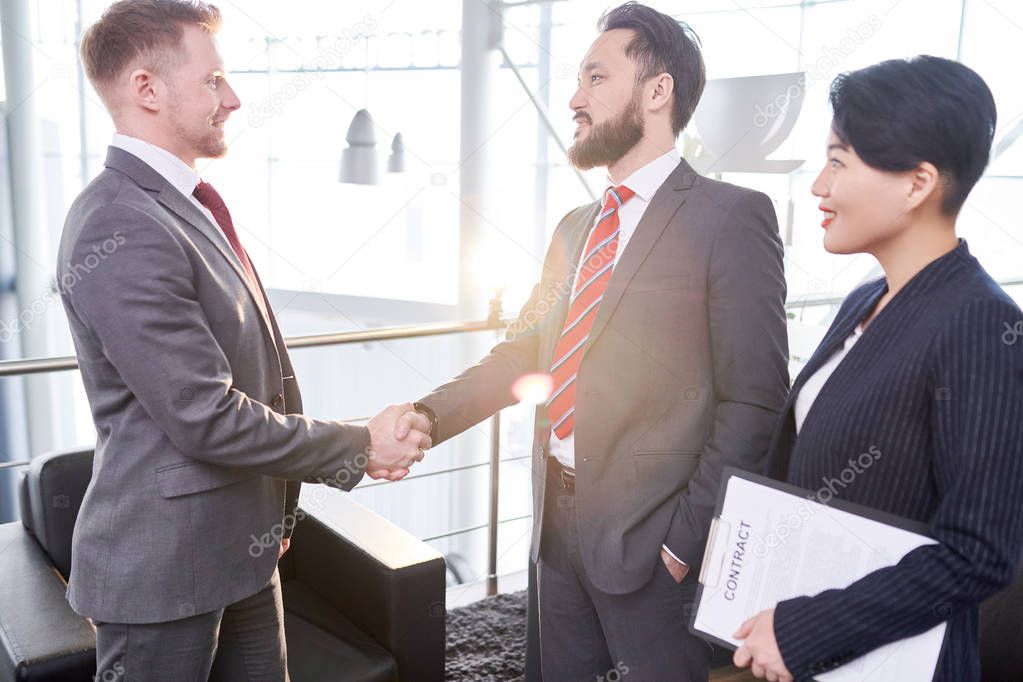 Profile view of confident entrepreneur wearing elegant suit greeting his business partner with firm handshake, his pretty assistant manager standing next to him, lens flare