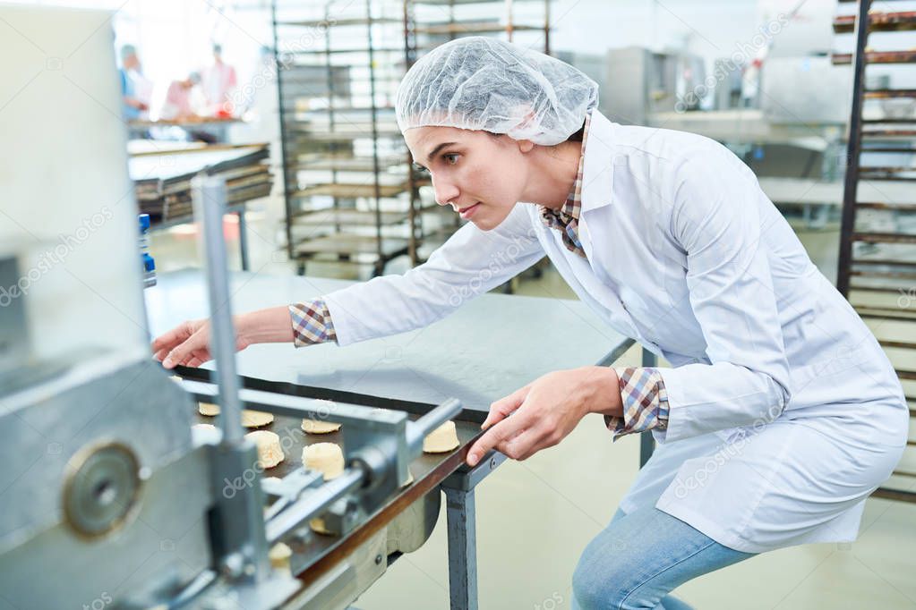 Confectionery factory employee in white coat holding baking sheet with uncooked pastry while working with machinery.