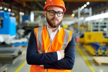 Modern sakallı işadamının hardhat ve yansıtıcı yelek giyen portresi, üretim atölyesinde güvenle poz verirken kameraya bakarak, kopya alanı