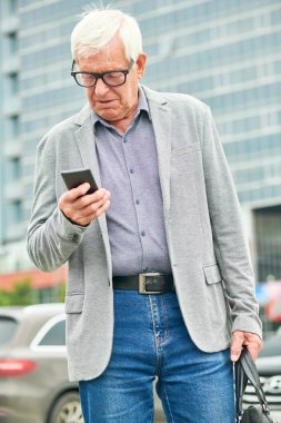 Şehir caddesinin bulanık arka planda dururken ceketli yaşlı adam akıllı telefona göz gezdiriyor