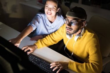 İki meslektaşın ekrana işaret eden ve gece geç saatlerde karanlık ofiste bilgisayar kullanırken projeyi tartışırken, önünde Orta Doğulu adama odaklanan yüksek açılı portresi