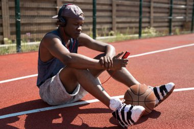 Modern Afrikalı basketboloyuncusunun açık hava sahasında yerde oturup akıllı telefon üzerinden müzik dinleyen tam uzunlukta portresi, kopya alanı