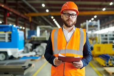 Modern sakallı işadamının hardhat ve yansıtıcı yelek giyen portresi, üretim atölyesinde poz verirken kameraya bakıyor, pano tutuyor, kopya alanı