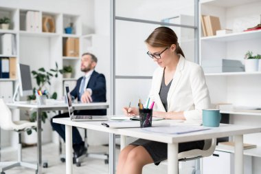 Beyaz ceket ve gözlük içerik meşgul çekici bayan masada oturan ve ofiste program planlama sırasında günlük notlar yapma