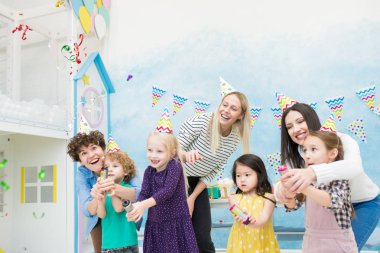 Neşeli kendinden geçmiş genç anne ve çocuklar renkli parti şapkaları mavi oyun odasında ayakta ve partide eğlenirken kraker patlama