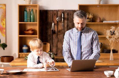 Beyaz gömlek ve kravat ciddi meşgul küçük oğlu masada duran ve baba olarak çalışırken gadget kullanarak, dizüstü bilgisayar ile çalışan sakallı baba