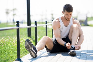 Parkta koşu pistinde otururken yakışıklı genç adam spor ayakkabıları bağlama tam uzunlukta portre, kopya alanı