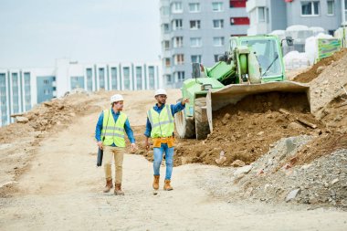 Kask lı ve yelekli iki adam kazıcının yanında yürüyor ve şantiyede yapılan inceleme sırasında mesafeyi işaret ediyor.