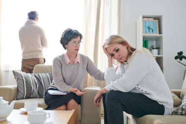 Olgun bayan psikologun genç kadının omzunu okşarken terapi seansında onu desteklerken kocası arka planda pencereden dışarı bakarken, ilişki zorlukları kavramını anlamak