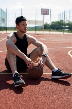 Açık basketbol sahasında katta oturan ve pensively uzak görünümlü yakışıklı kas adam tam uzunlukta portre, kopya alanı
