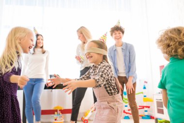 Oyun sırasında arkadaş yakalarken parti şapka gesturing elleri pozitif heyecanlı gözleri bağlı kız, neşeli çocuklar gözleri bağlı kız kaçan