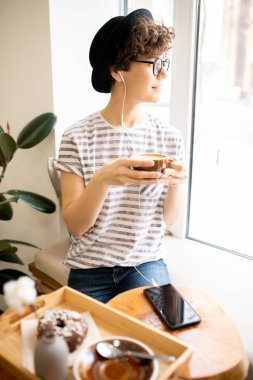 Şapka ve gözlük pensive güzel genç bayan kafede pencere eşik üzerinde oturan ve pencereden dışarı bakarken ve kulaklık müzik dinlerken taze demlenmiş kahve içme.