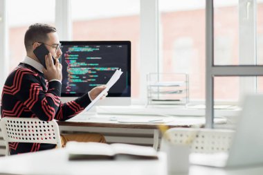 Ciddi meşgul yakışıklı sakallı programcı kazak bilgisayar ile masada oturan ve belge metnini açıklarken telefonla müşteri ile konuşurken
