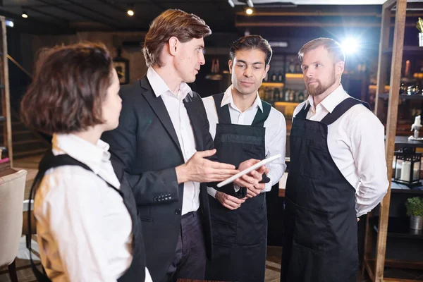 Serious confident restaurant manager  using digital tablet and pointing at screen while discussing seating chart with waiters before banquet