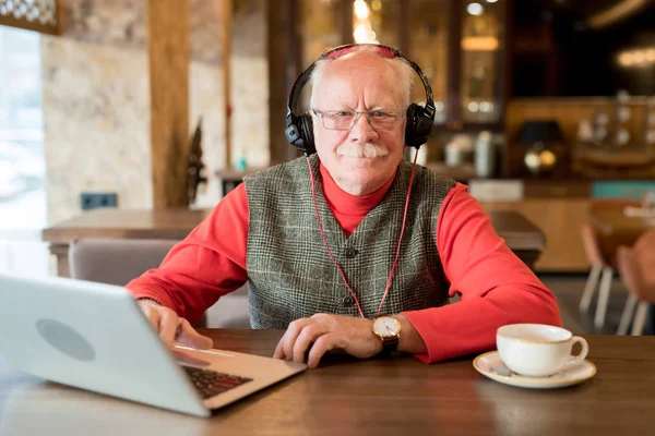 Content handsome modern elderly man in headphones wearing stylish waistcoat and red turtleneck sitting at table and looking at camera while resting in cafe