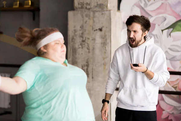 Portrait of angry coach shouting at  obese young woman in fitness club, copy space