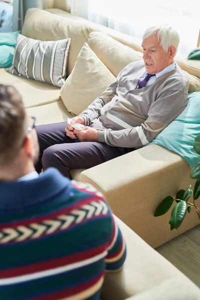 Serious handsome elderly man in cardigan sitting on sofa and holding napkin while explaining his feeling to psychiatrist during therapy session