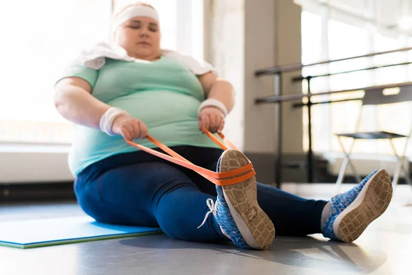Full length portrait of young obese woman exercising with elastic band in fitness club, focus on foreground