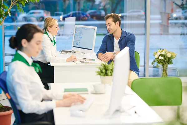 Cheerful attractive bank sales consultant in corporate outfit sitting at table with computer and showing magazine with bank products to customer while working with him in office.