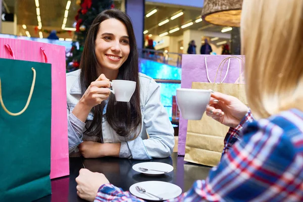 Smiling young woman resting in a cafe and drinking coffee with her friend after shopping