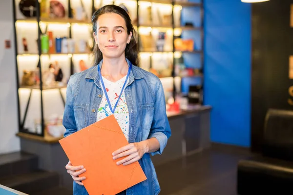 Content confident female manager of forum wearing badge on neck holding red clipboard and standing against shelf with souvenir products